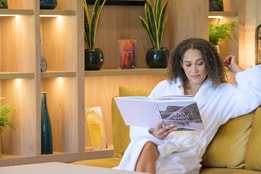 A woman in a robe sits comfortably on a couch, absorbed in her book, enjoying the peaceful ambiance of Heythrop Park Spa.
