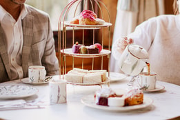 Tea being poured from a teapot into a teacup next to a tiered stand of sandwiches, cakes, and macarons, with plates of scones and jam arranged on the table.
