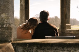 Two people sit closely together on a bench inside a stone structure with tall columns, facing an open landscape of grass and distant trees. One person leans their head against the other’s shoulder. Warm sunlight backlights the scene, creating a soft, quiet atmosphere.