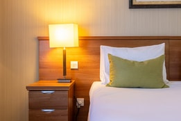 Close-up of a hotel bed with bedside table, wooden headboard and soft lighting.
