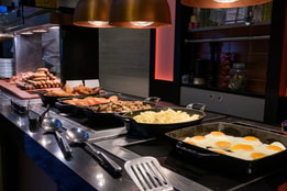 Hot breakfast buffet with trays of fried eggs, scrambled eggs, mushrooms, tomatoes, sausages, and hash browns under heat lamps.