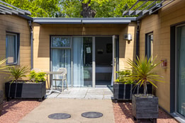 A modern lodge exterior with sliding glass doors opening onto a small patio, featuring a table, chairs, and large planters with greenery.
