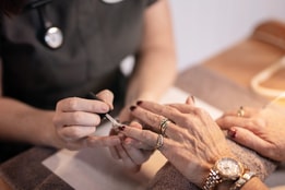 Close-up of a manicure in progress, with a technician applying dark nail polish to a client’s fingernails while hands rest on a towel-covered surface, surrounded by soft, warm lighting.