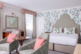 A spacious bedroom with a large bed against patterned wallpaper, paired with two green armchairs and a round table in the foreground.