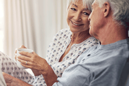 An elderly couple sits closely together in bed, sharing a warm drink, radiating love and companionship in a cozy setting.