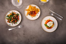 Overhead view of three small plates with colourful dishes, including salad and flatbread with dip, alongside glasses of iced drinks with lemon on a stone table.