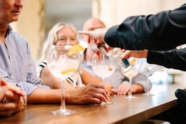 A bartender pours a drink into a large cocktail glass, surrounded by friends enjoying drinks at a stylish bar.