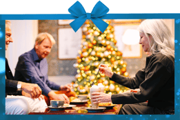 Three people enjoying hot drinks in front of a christmas tree with a bow border