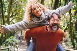 A joyful moment in a forest: a husband giving a piggyback ride to his wife, with arms raised, surrounded by lush greenery., on a Warner break.