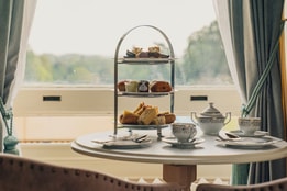 A charming afternoon tea set with an elegant three-tier stand filled with pastries, beside fine china cups and a teapot, against a scenic window view.