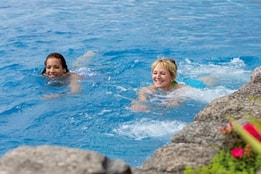 Two women swimming in a vibrant blue pool, with rocky edges and colorful flowers nearby, creating a lively summer atmosphere.