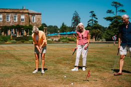 Three seniors play golf on a sunlit lawn near a historic building, focused on their shots with flags marking the holes.