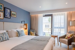 New Deluxe room at Heythrop Park Hotel with a king-sized bed, plush pillows, decorative art, and chairs beside a window overlooking a garden patio.