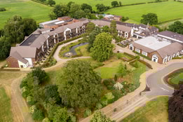 Aerial view of Alvaston Hall hotel  - a  landscaped resort with buildings arranged around gardens, pathways, and a pond, surrounded by lush green fields.