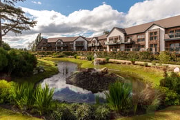 Serene landscape at Alvaston Hall featuring modern accommodations beside a tranquil pond with a fountain, surrounded by lush greenery and blue skies.