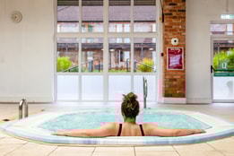 A woman relaxes the bubble pool at Alvaston Hall Hotel, facing large windows that overlook a green garden area. A clock is visible on the wall.