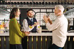 Two guests smiling at each other as a barman makes their cocktails