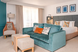 Spacious contemporary hotel room with a king-size bed, teal sofa, upholstered armchair and footstool. Large windows with floor-length curtains provide natural light, while coordinated furnishings and colourful accent cushions create a stylish and welcoming setting.