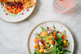 Overhead view of a fresh salad with roasted squash alongside flatbread with dip, served with a pink iced drink garnished with strawberries on a marble table.