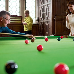 Person taking a shot on a snooker table in a wood-panelled room, with two other people standing nearby. Red and coloured balls are spread across the green baize under natural light from large leaded windows.