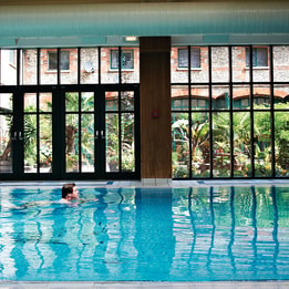 Person swimming in an indoor pool beside floor-to-ceiling windows overlooking a landscaped courtyard. Reflections shimmer across the water, creating a calm and relaxing leisure environment.