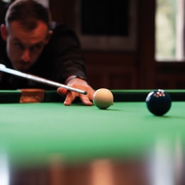 Person leaning over a snooker table lining up a shot, cue aimed at the white ball with coloured balls nearby, set against a dimly lit bar or games room background.