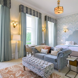 Spacious manor house bedroom at Heythrop Park featuring a bed, chair, lamp, and a large window for natural light.