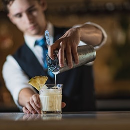A stylish barman in a suit serves a drink into a glass at the bar of the Runnymede on Thames hotel.