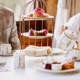 Tea being poured from a teapot into a teacup next to a tiered stand of sandwiches, cakes, and macarons, with plates of scones and jam arranged on the table.