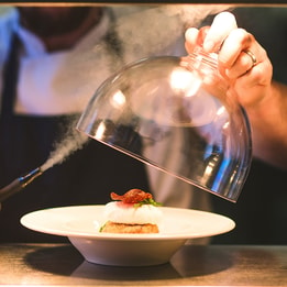 Chef lifting a glass cloche as smoke escapes over a plated smoked fishcake topped with a poached egg and crisp garnish, creating a dramatic presentation.