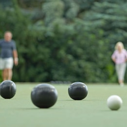 Close-up of black and white bowls on a bowling green, with players blurred in the background and trees surrounding the outdoor playing area.