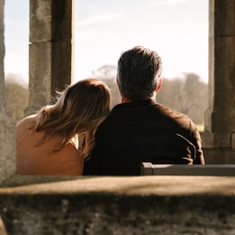 Two people sit closely together on a bench inside a stone structure with tall columns, facing an open landscape of grass and distant trees. One person leans their head against the other’s shoulder. Warm sunlight backlights the scene, creating a soft, quiet atmosphere.