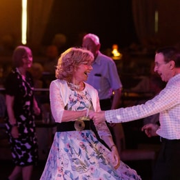 A woman in a floral dress dances with a partner in the warmly lit entertainment lounge at Alvaston Hotel, filled with seated guests.