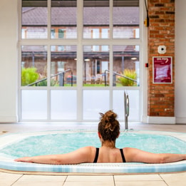A woman relaxes the bubble pool at Alvaston Hall Hotel, facing large windows that overlook a green garden area. A clock is visible on the wall