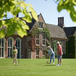 People playing croquet on a large lawn in front of a historic ivy-covered manor house, framed by green tree leaves in the foreground on a bright summer day.