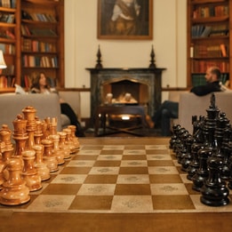 Chess set arranged on a wooden table in a traditional library lounge, with bookshelves, armchairs and a fireplace creating a warm, welcoming setting.