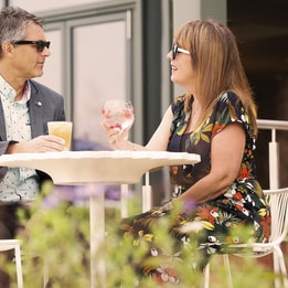 Two people seated at a white table outdoors at Studley Castle, holding drinks and engaging in conversation with plants in foreground.