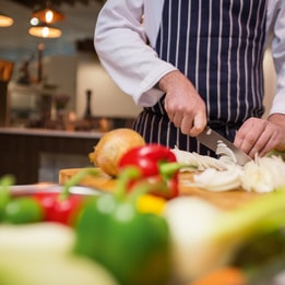 Chef slicing an onion on a wooden chopping board, surrounded by fresh vegetables including bell peppers and leeks on a kitchen counter.
