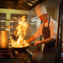 Chef in a professional kitchen cooking with a pan over open flames on a gas stove, with a large pot beside it and industrial kitchen equipment in the background.