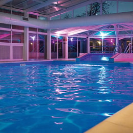 Indoor spa pool illuminated with pink and blue ambient lighting beneath a glass roof
