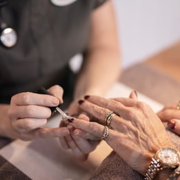 Close-up of a manicure in progress, with a technician applying dark nail polish to a client’s fingernails while hands rest on a towel-covered surface, surrounded by soft, warm lighting.