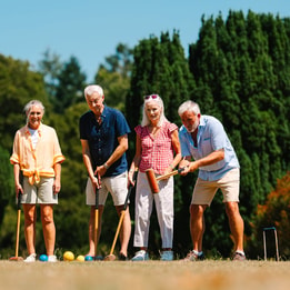 A group of six individuals wearing casual summer clothing stand on a grassy field, preparing to play croquet against a sunny backdrop.