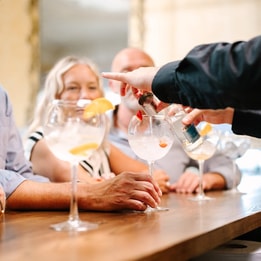 A bartender pours a drink into a large cocktail glass, surrounded by friends enjoying drinks at a stylish bar.