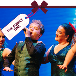 Festive stage performance featuring a singer holding a microphone, surrounded by performers in sequinned outfits under bright blue lighting. A gift tag tied with a red ribbon reads ‘The Bubbly One’.