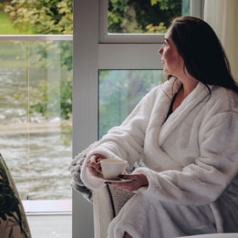 A woman in a robe sits comfortably in a chair, gazing out the window, bathed in soft natural light.