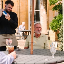 A waiter in a black shirt shakes a cocktail shaker at an outdoor table, while guests enjoy drinks under a sunny sky a Cricket St. Thomas