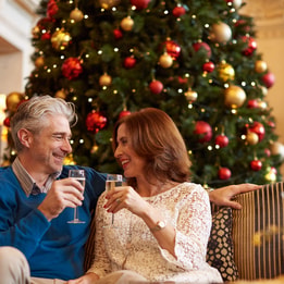 A couple enjoying a drink together in front of a Christmas tree and smiling at one another in the Great Hall at Thoresby Hall