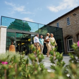 Four people walk towards a modern glass entrance, surrounded by greenery and flowers on a sunny day.