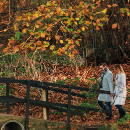 A couple walks hand-in-hand over a wooden bridge surrounded by autumn foliage and fallen leaves.