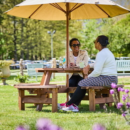 Two women sit at a wooden picnic table under a large umbrella, surrounded by lush greenery and vibrant flowers at Sinah Warren